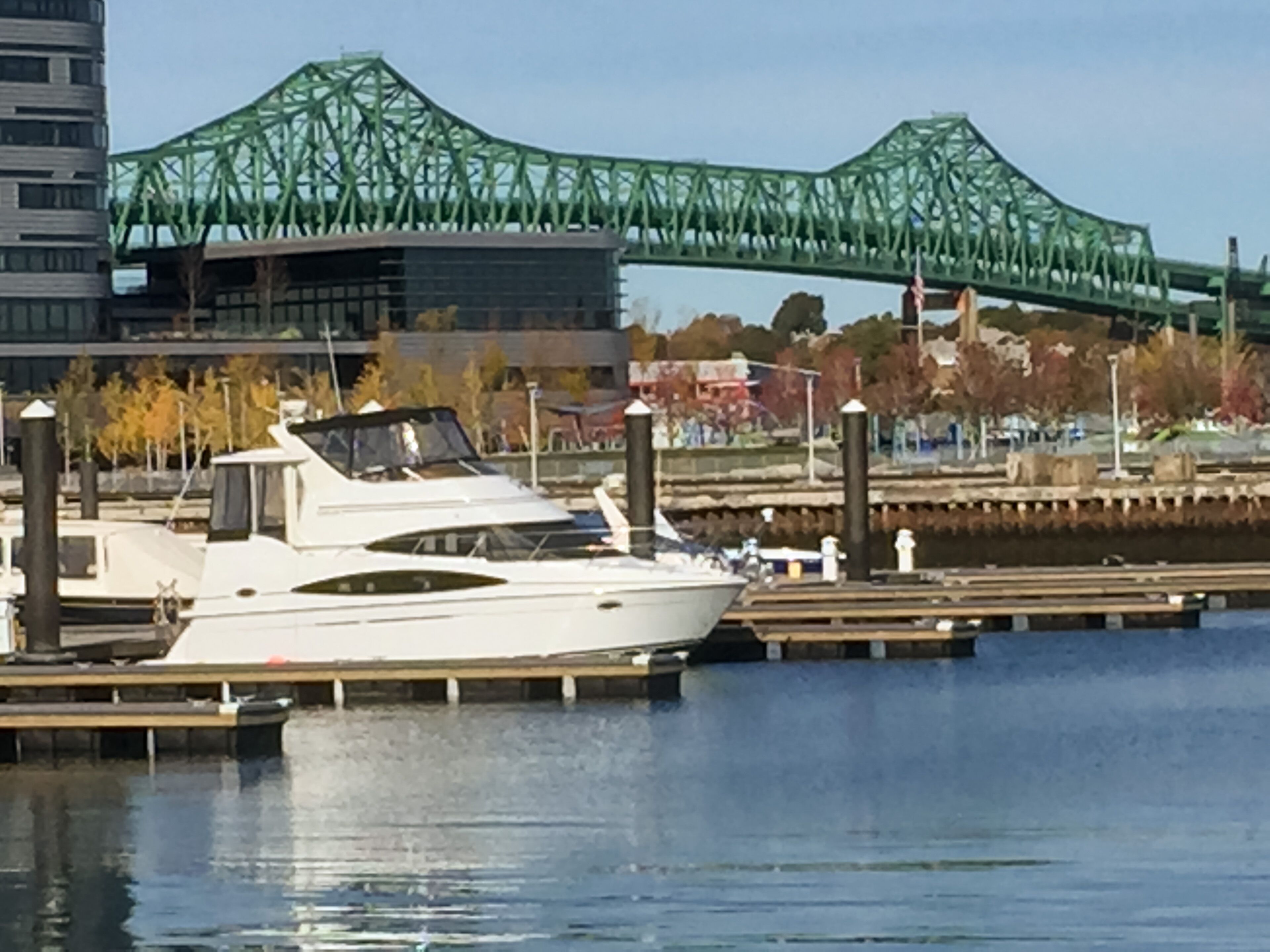 Un port de plaisance où un yacht blanc est amarré à un quai en bois, un pont vert en arrière-plan et un paysage urbain avec des arbres aux couleurs automnales.