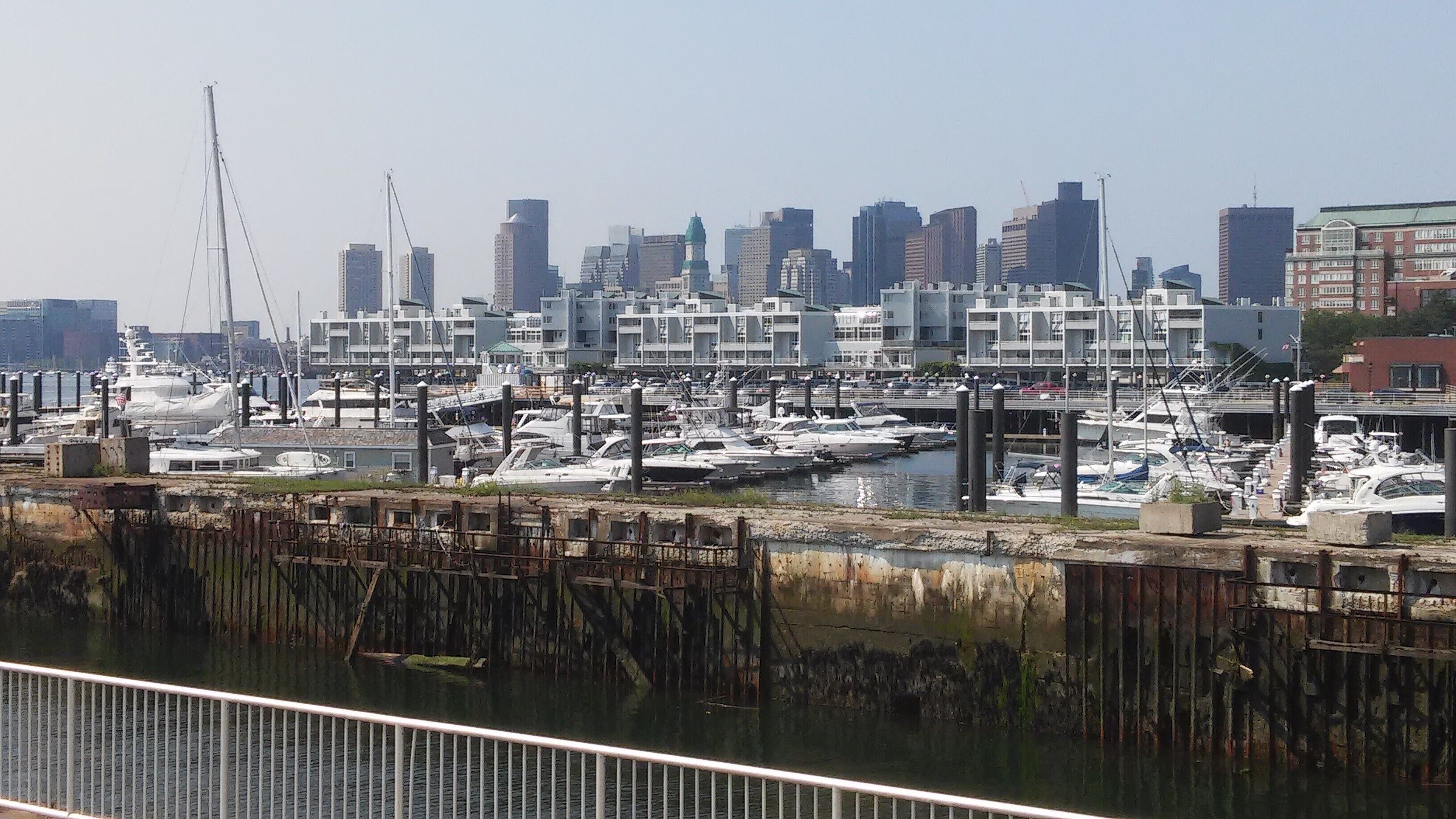 Un puerto deportivo con numerosas embarcaciones amarradas, un horizonte urbano al fondo y un muelle desgastado en primer plano.