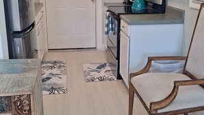 Cocina compacta con muebles blancos, un refrigerador, un horno y un microondas. Hay una zona de comedor con una silla de madera y una mesa.