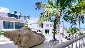 A balcony with a large rock, palm trees, and a view of a white building with balconies.