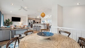 A modern living room with a round wooden dining table, a TV mounted on the wall, and a kitchen in the background. - Pet-Friendly, Close to beach access & town, Large - 2100 sq ft! Afternoon Delight! (Folly Beach)