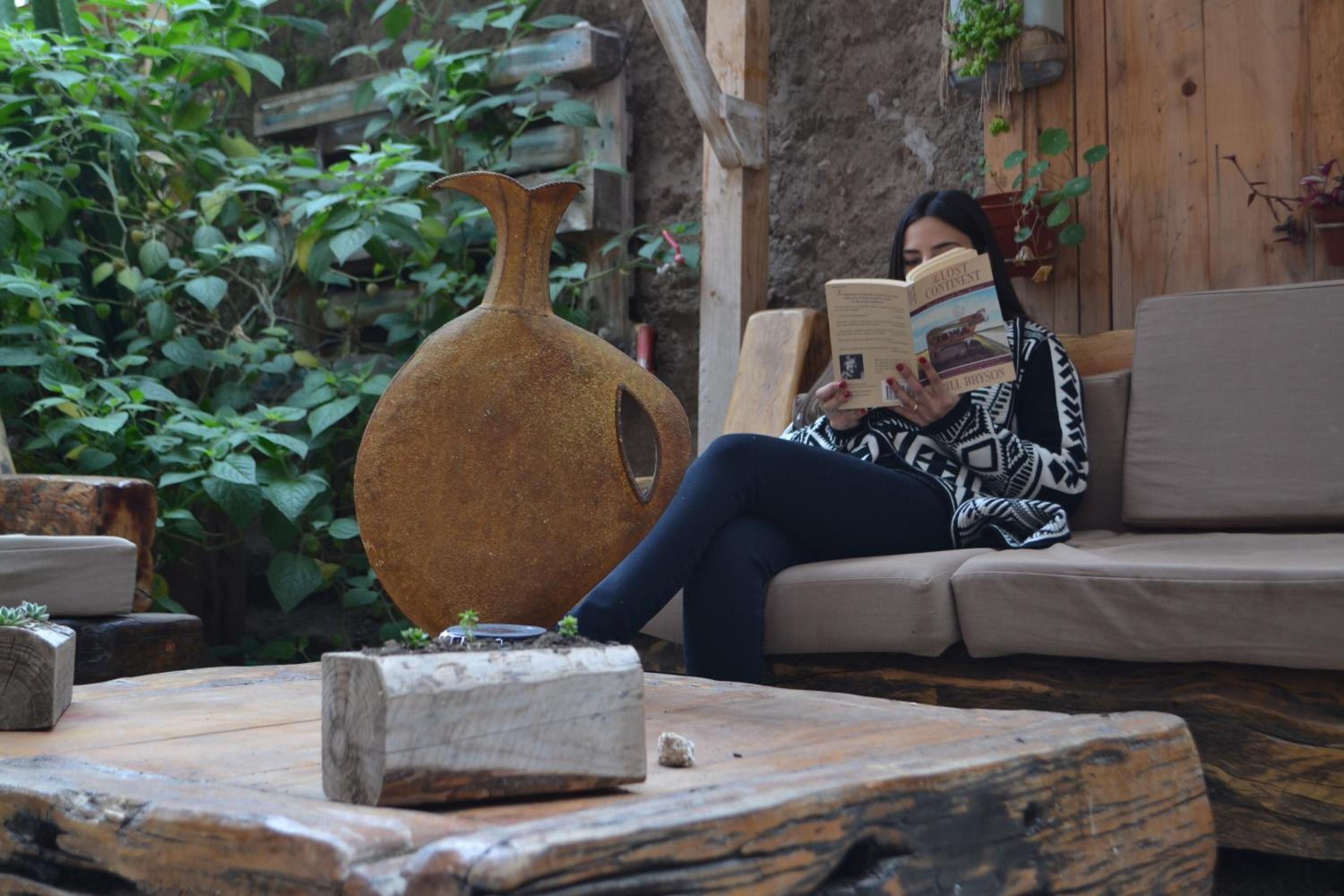 A person sitting on a sofa reading a book in a rustic setting with wooden furniture and greenery.