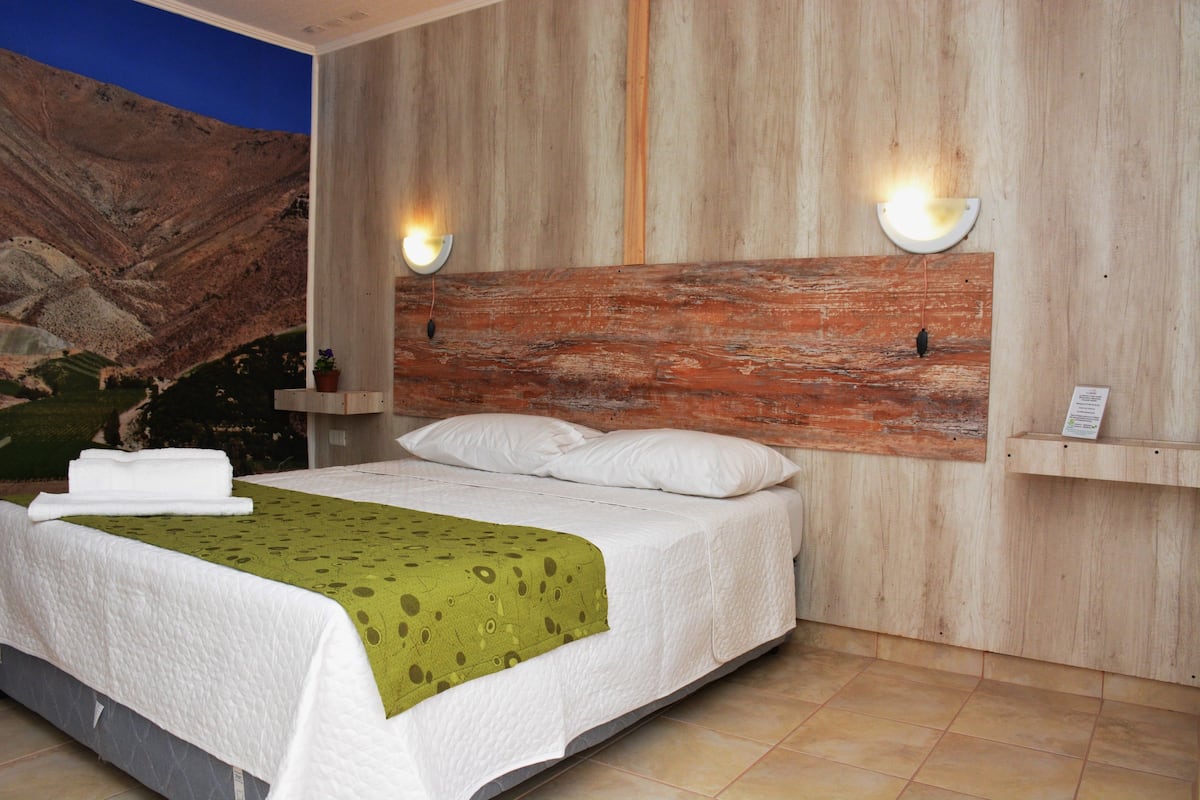 Ein Schlafzimmer mit einem Holzkopfende, einem Bett mit weißen Leinen, zwei Wandleuchten und einer Bergmalerei an der Wand.