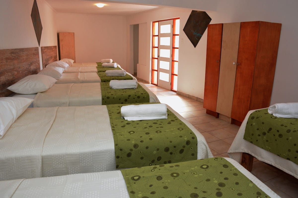 Ein Hotelzimmer mit vier Einzelbetten, jeweils mit grüner Bettdecke und weißen Kissen. Ein Holzschrank und eine Tür zu einem hellen Raum sind ebenfalls vorhanden.
