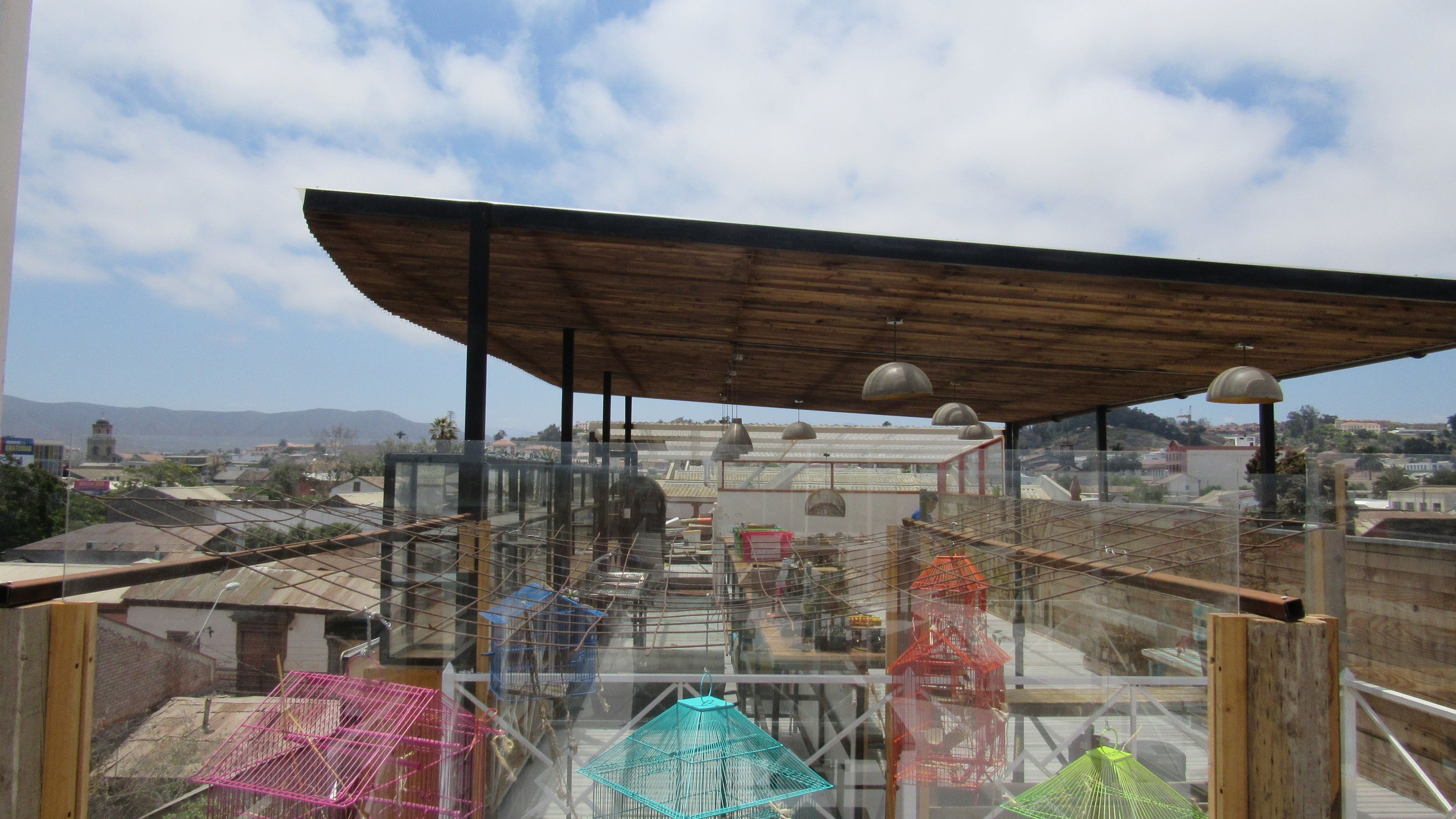 A rooftop terrace with a wooden pergola, metal birdcages, and a view of the cityscape.