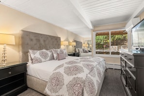 Ein Schlafzimmer mit einem großen Bett, einem Nachttisch, einer Kommode und einem Fenster mit Blick auf Grünflächen.