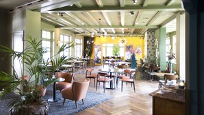 A spacious dining area with multiple tables and chairs, large windows, and potted plants. - De Kastanjehof (Lage Vuursche)