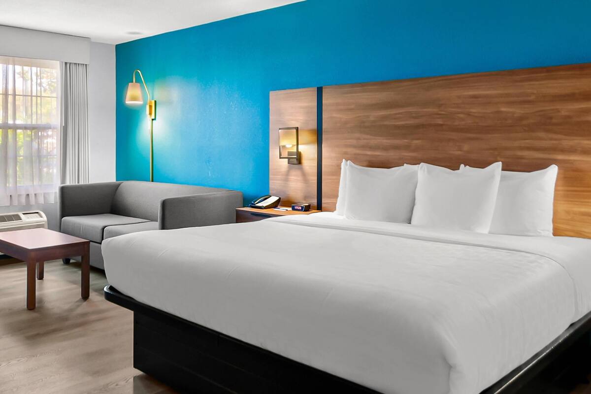 A hotel room with a large bed, a grey sofa, a wooden headboard, a bedside table with a lamp, and a window with curtains.