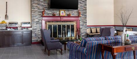 A living room with a fireplace, striped sofas, a TV mounted on the wall, and a kitchen area with appliances.