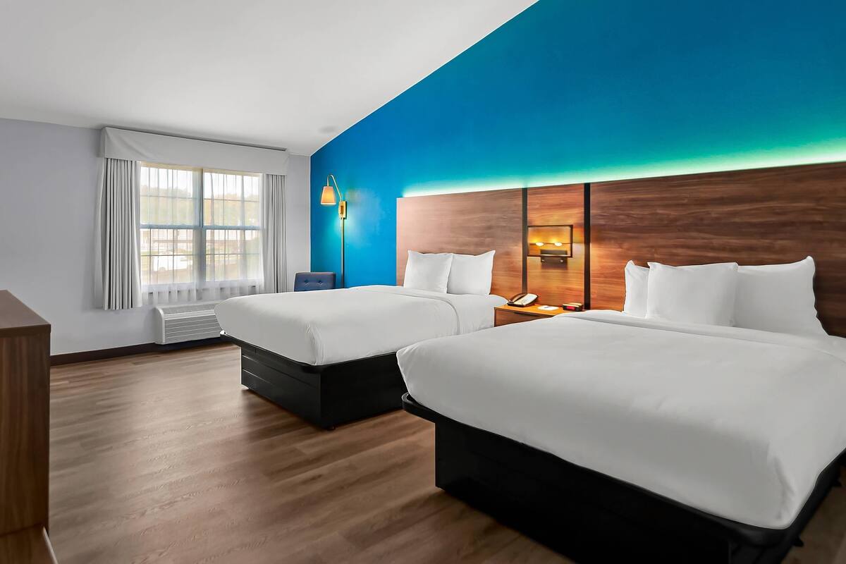 A hotel room with two beds, a large window with curtains, a bedside table with a lamp, and a wall-mounted phone.