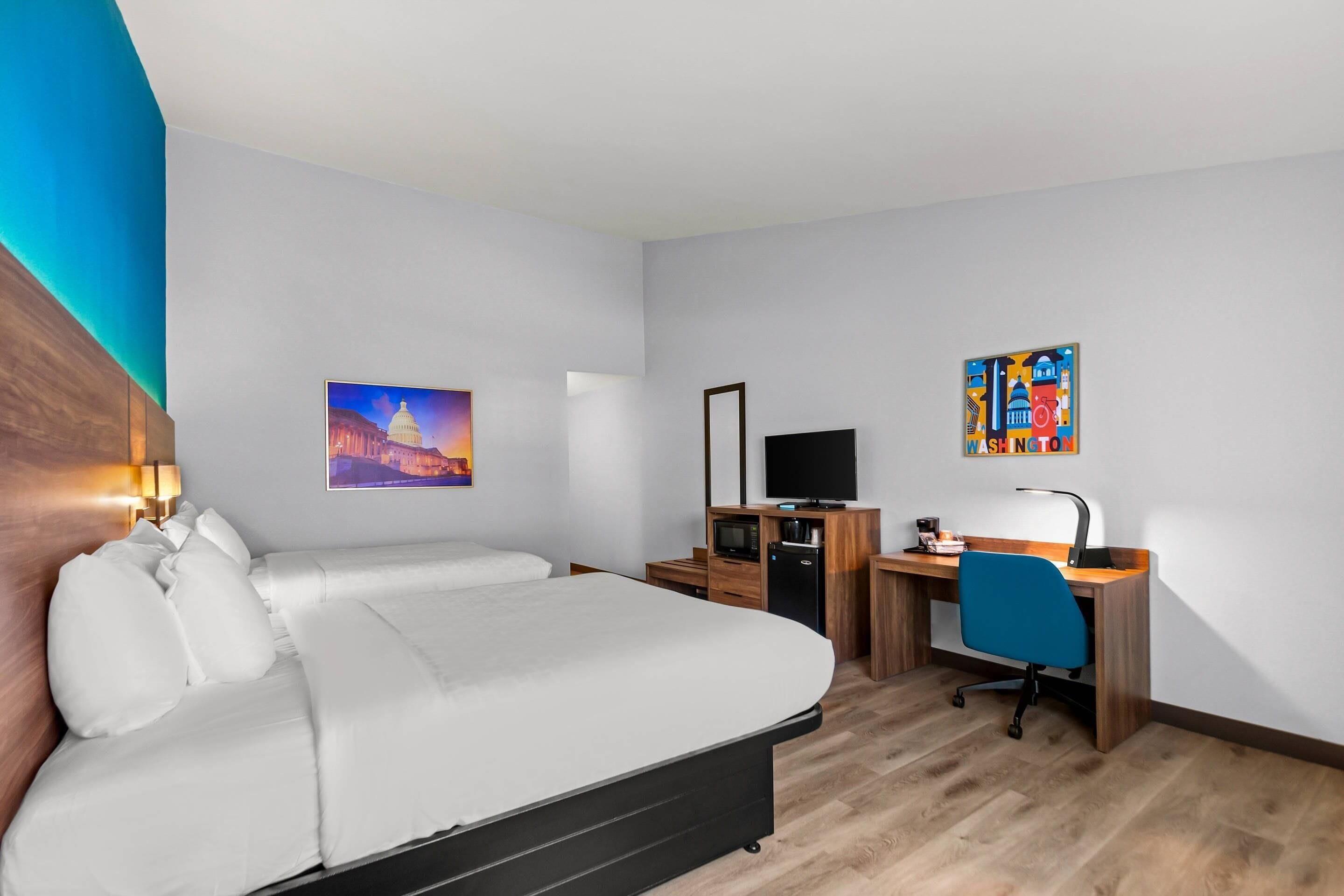 A hotel room with a bed, desk, TV, and artwork.