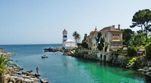 Escena costera con un faro, una casa grande con jardín y aguas azules cristalinas.