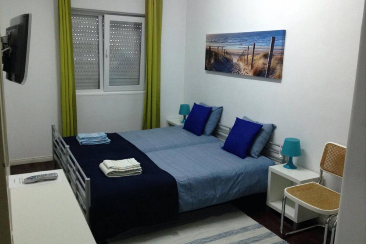 Un dormitorio con cama, dos mesitas de noche, una silla, un cuadro y una ventana con cortinas.