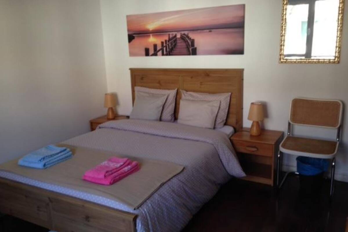 Un dormitorio con cabecera de madera, cama con manta gris, dos almohadas, una mesita de noche con lámpara, una silla y un cuadro de un atardecer sobre un muelle.