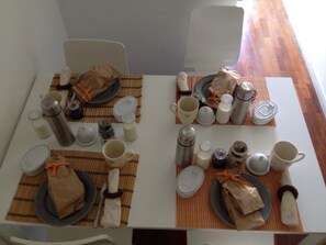 Una mesa de comedor dispuesta con platos, cubiertos y diferentes vasos, incluyendo termos y tazas, sobre un mantel a cuadros.