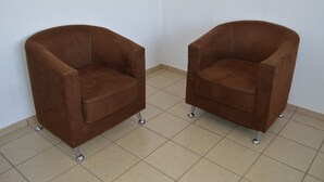 Twee bruine fauteuils met metalen poten, staand op een betegelde vloer tegen een effen muur.