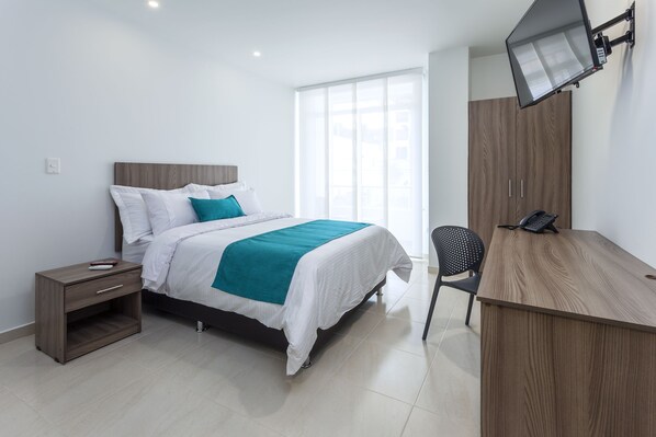 A modern bedroom with a bed, bedside table, desk, chair, and a TV mounted on the wall.