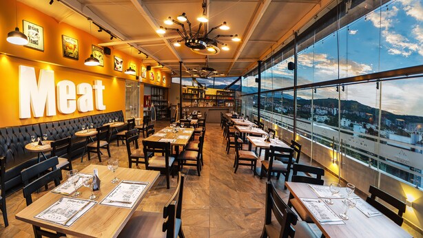 A restaurant with a large 'Meat' sign, wooden tables, and chairs, and a view of the city through large windows. - Torca Hotel (Cali)