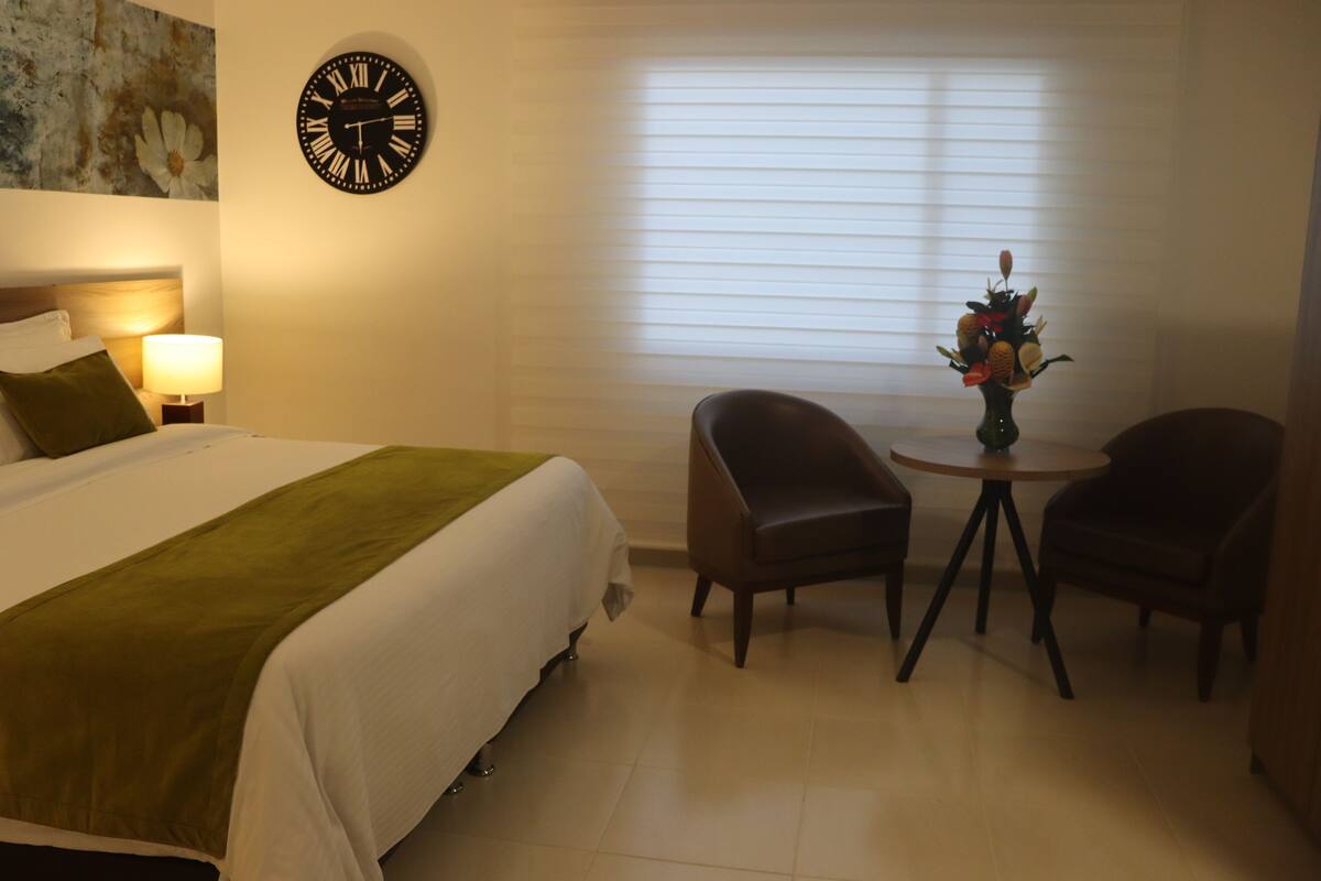 A hotel room with a bed, a clock, a lamp, two chairs, and a small table with a vase of flowers.