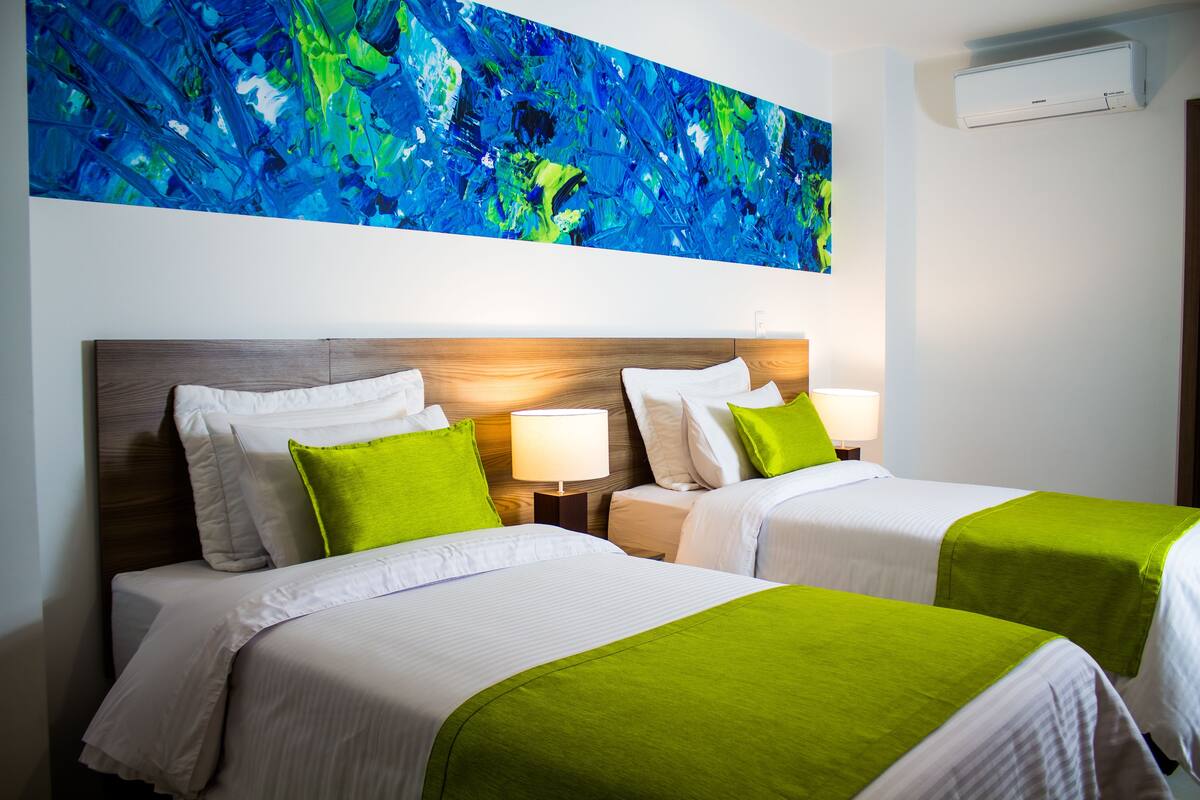 A hotel room with two beds, each with white bedding and green accents, a large abstract painting on the wall, and an air conditioning unit.