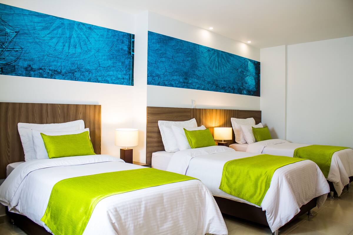 A hotel room with two beds, each with white bedding and green throw pillows. There are two wall-mounted lamps on either side of the beds.