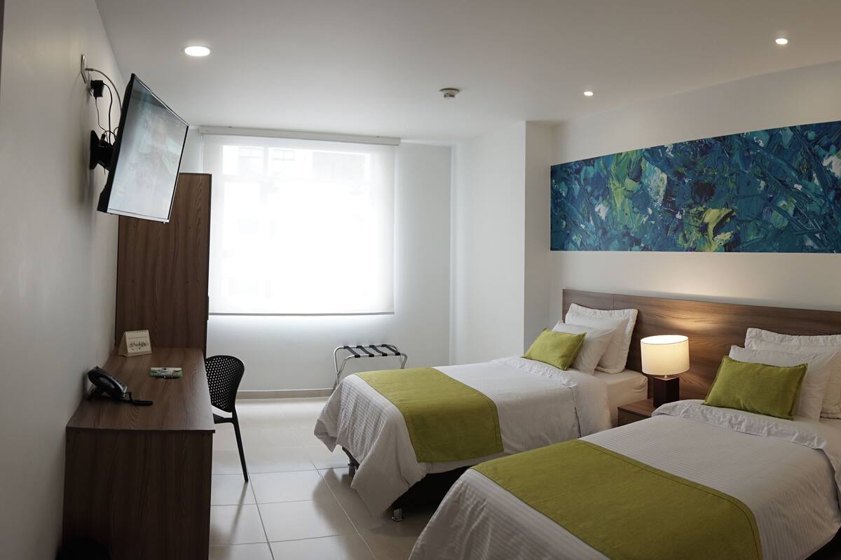 A hotel room with two beds, a desk, a chair, a TV, and a large abstract painting on the wall.