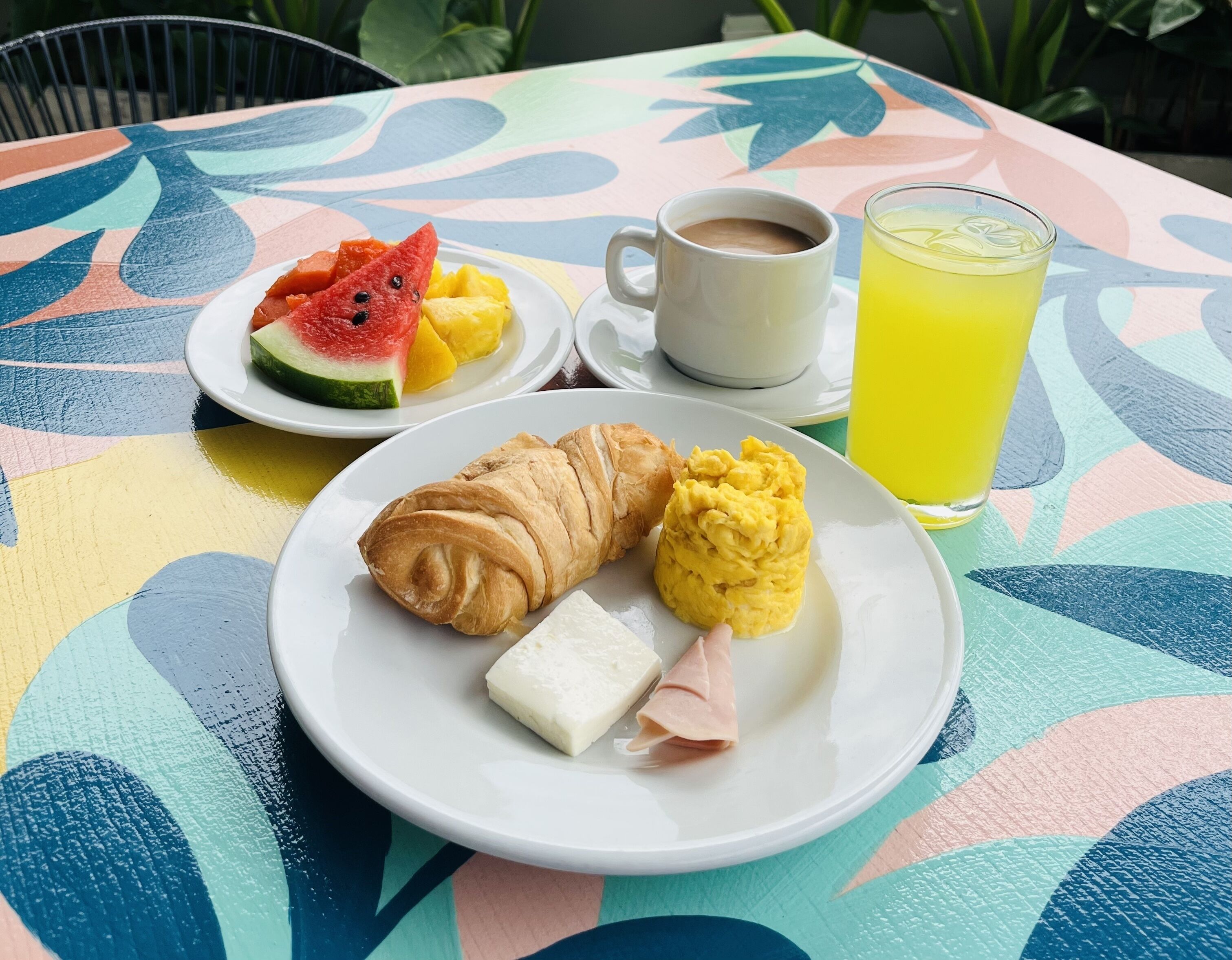 A table with a colorful tablecloth, a plate with croissant, cheese, and scrambled eggs, a plate with watermelon and mango, a cup of coffee, and a glass of yellow drink.