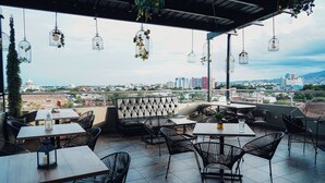 An outdoor seating area with tables and chairs, a cushioned bench, and hanging birdcages with plants. - Torca Hotel (Cali)