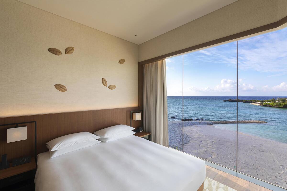 A hotel room with a large bed, a nightstand, a wall clock, and a view of the sea.