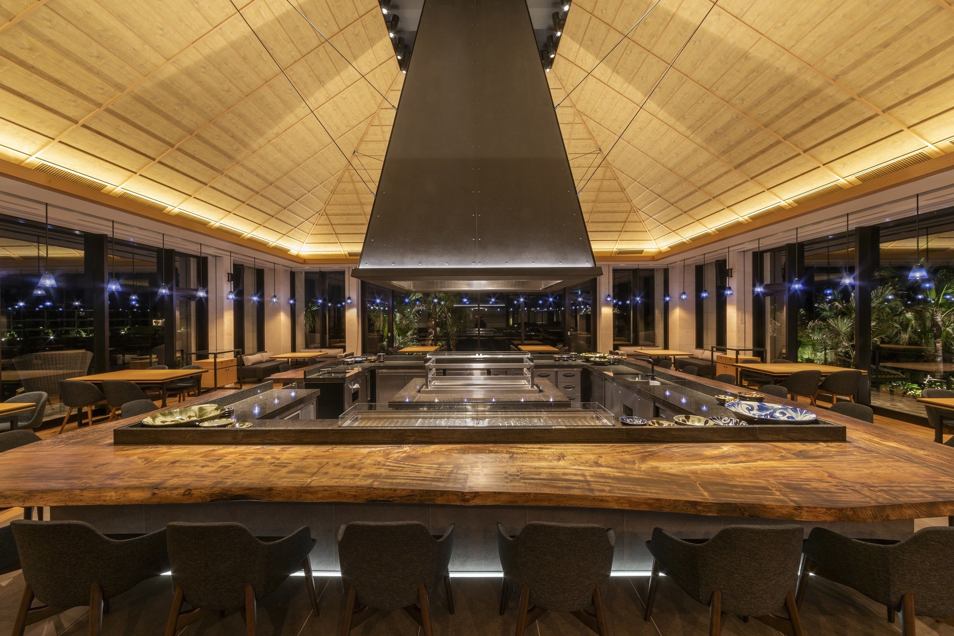 Un restaurant moderne doté d’un bar central, de plusieurs espaces de détente et d’un plafond haut orné de panneaux en bois.