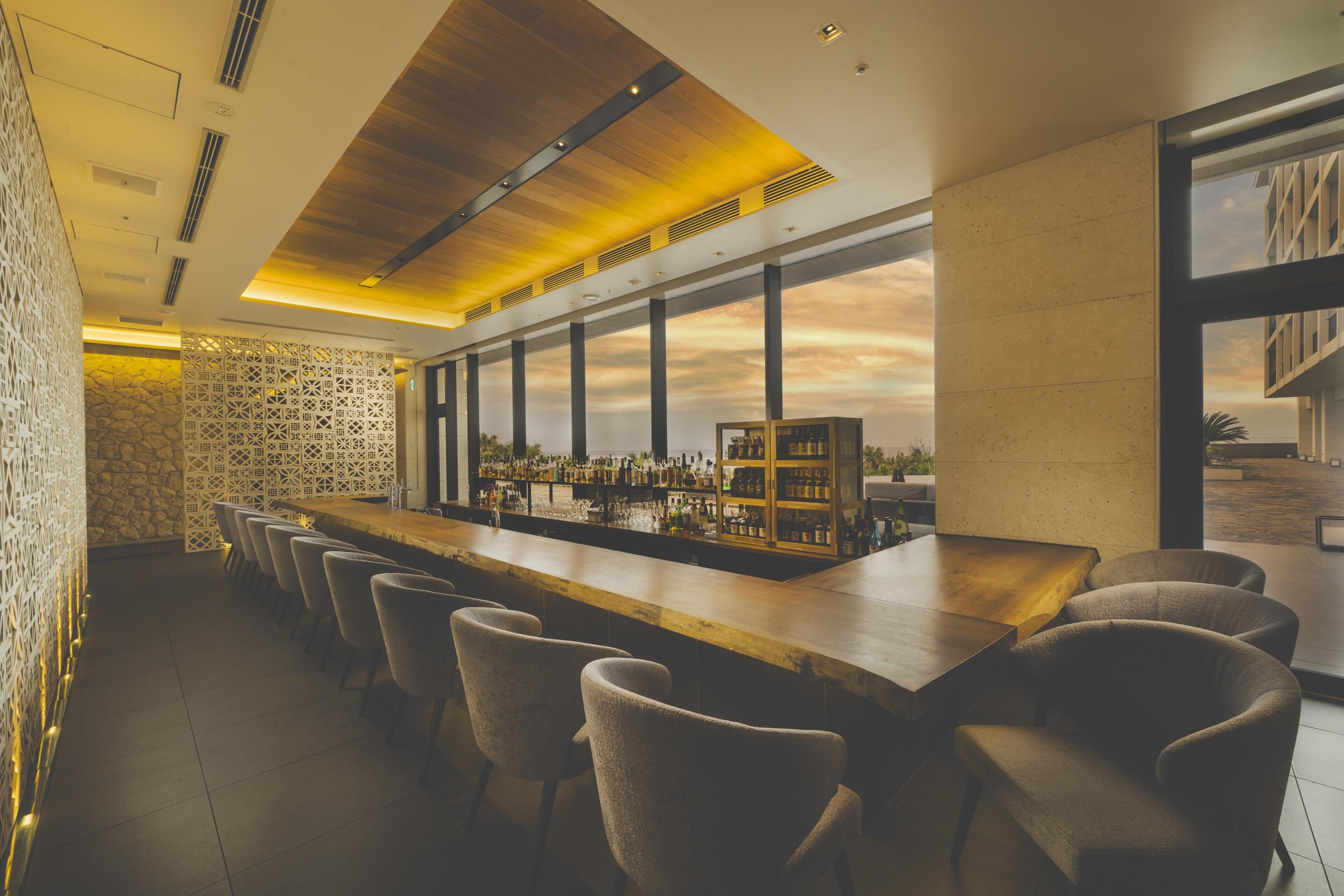 A bar area with a long counter, multiple bar stools, and a view of the sky through large windows.