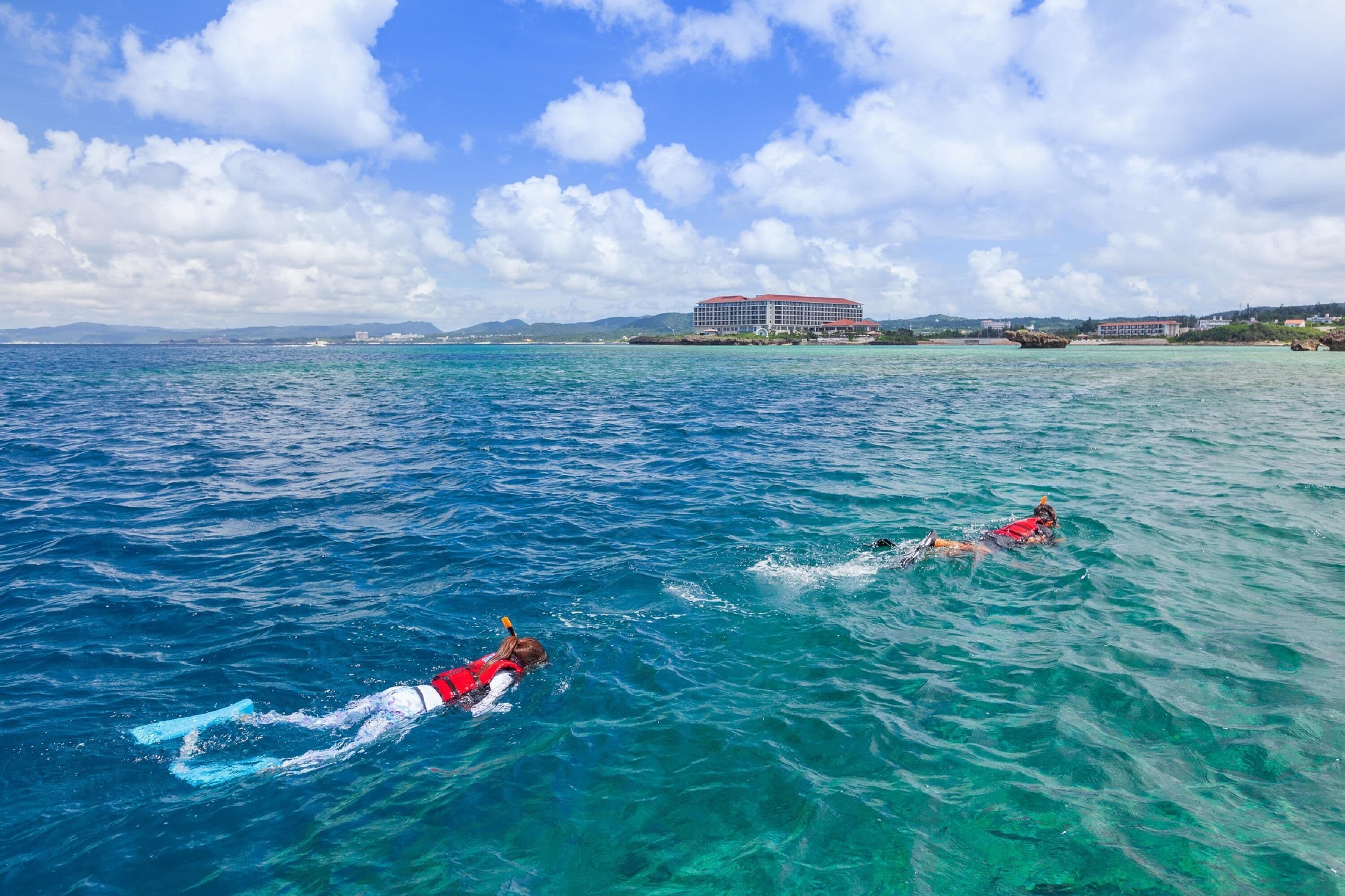 Two people swimming in clear blue water with a building in the background.