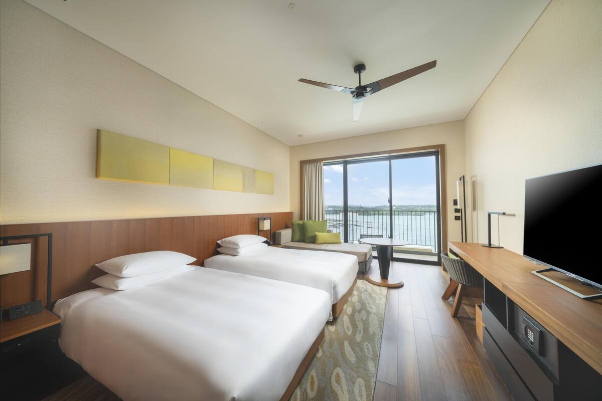 A hotel room with two beds, a TV, a balcony with a view, and a ceiling fan.