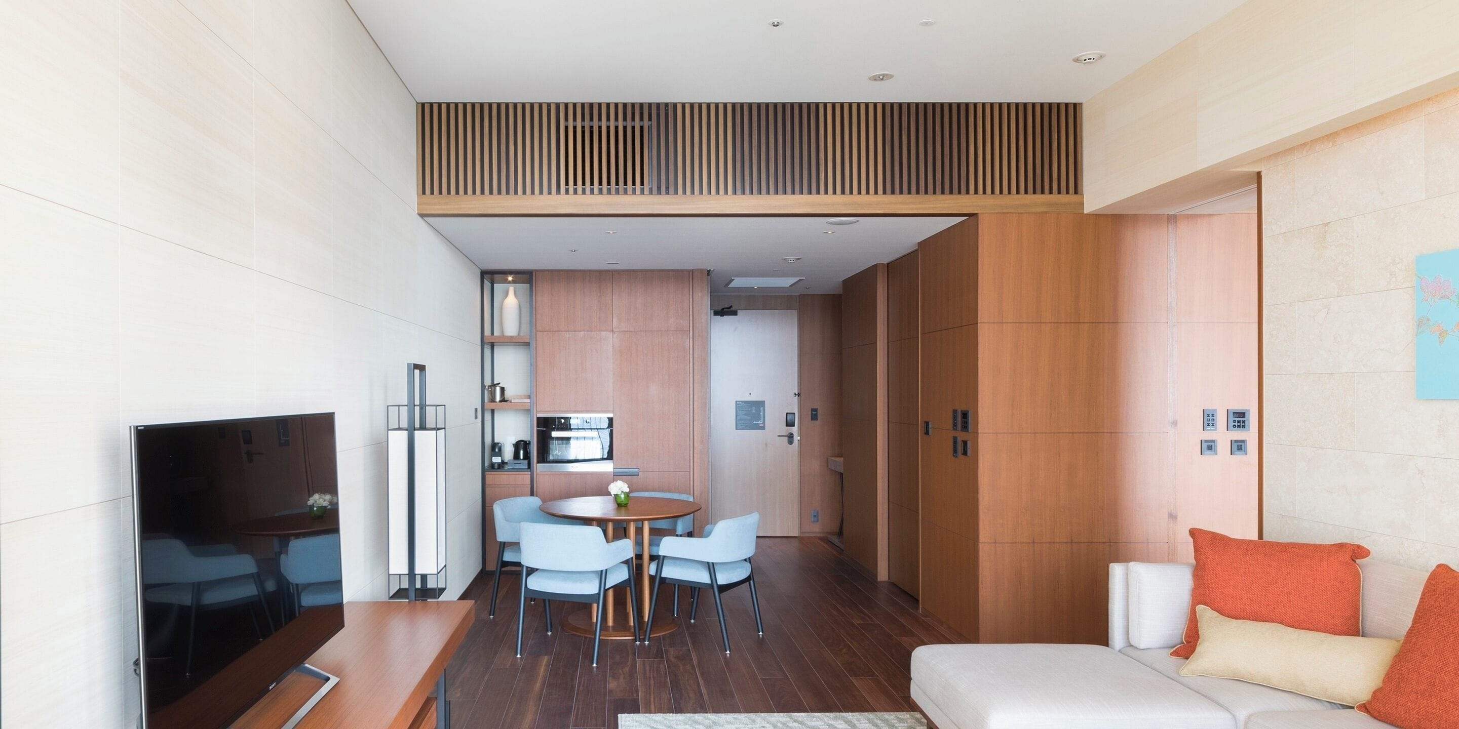A modern hotel room with a dining area, kitchenette, and a sofa with orange pillows.