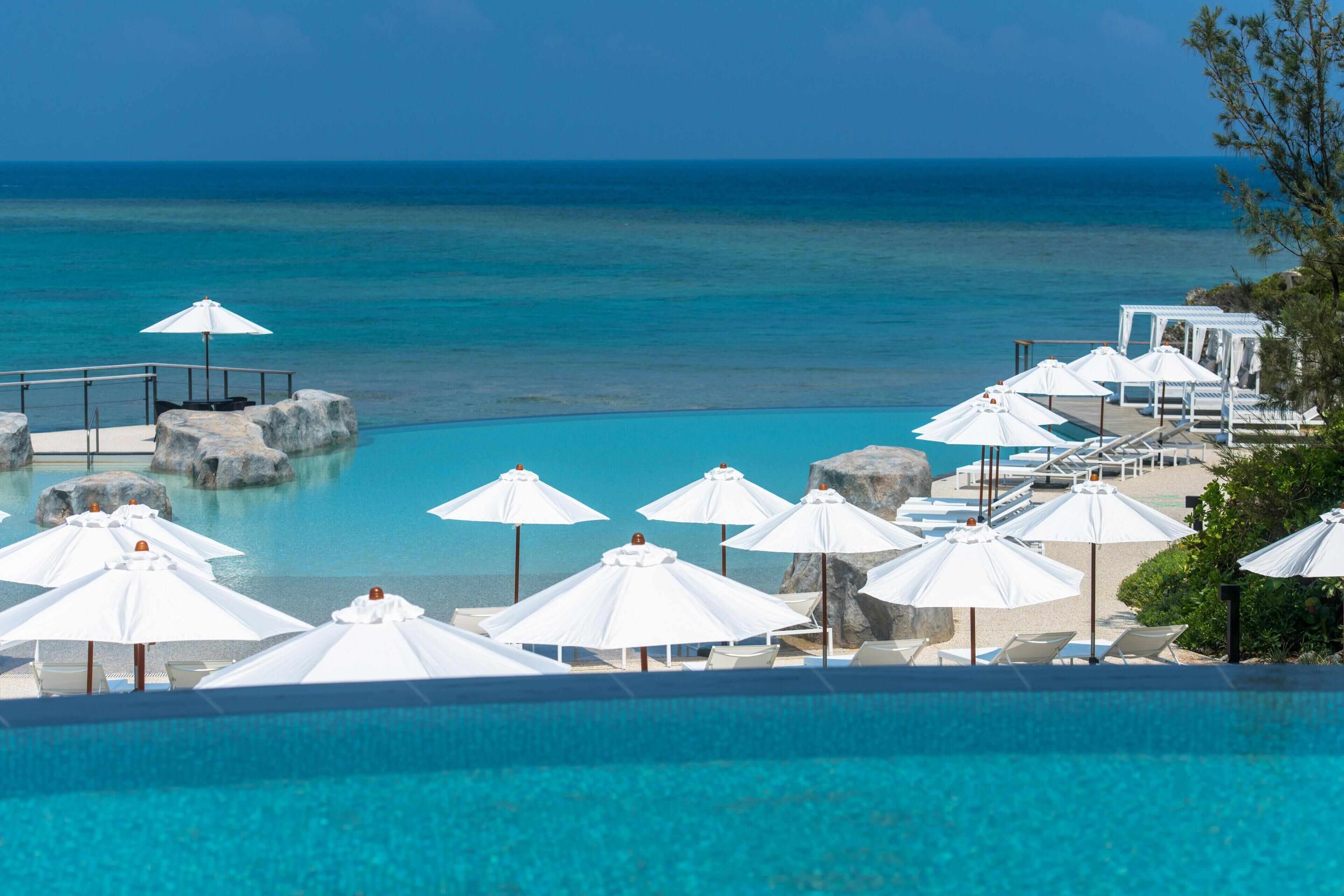 A pool area with umbrellas and lounge chairs, overlooking a clear blue sea.