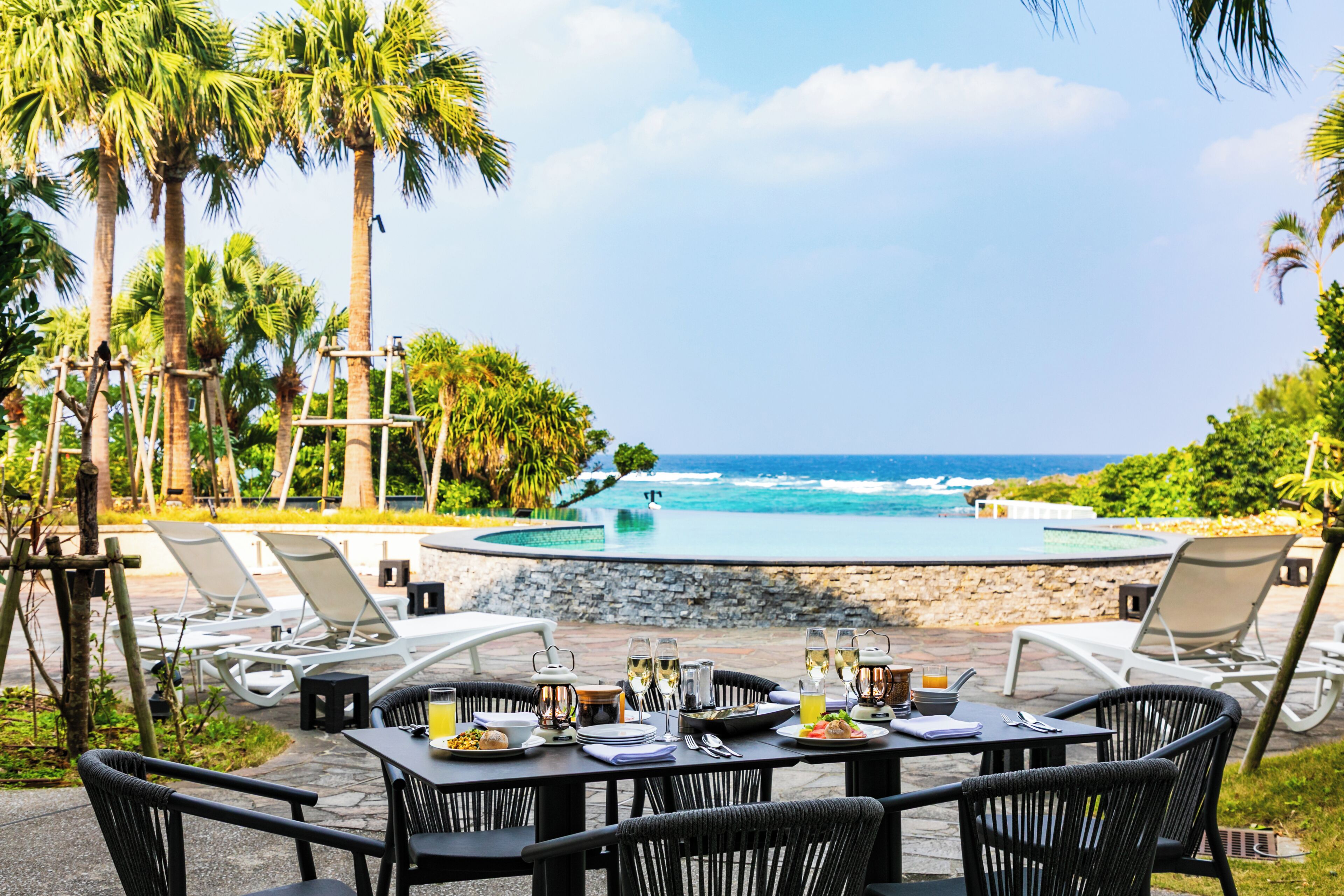 A poolside dining area with lounge chairs, a table set for a meal, and a view of the ocean.