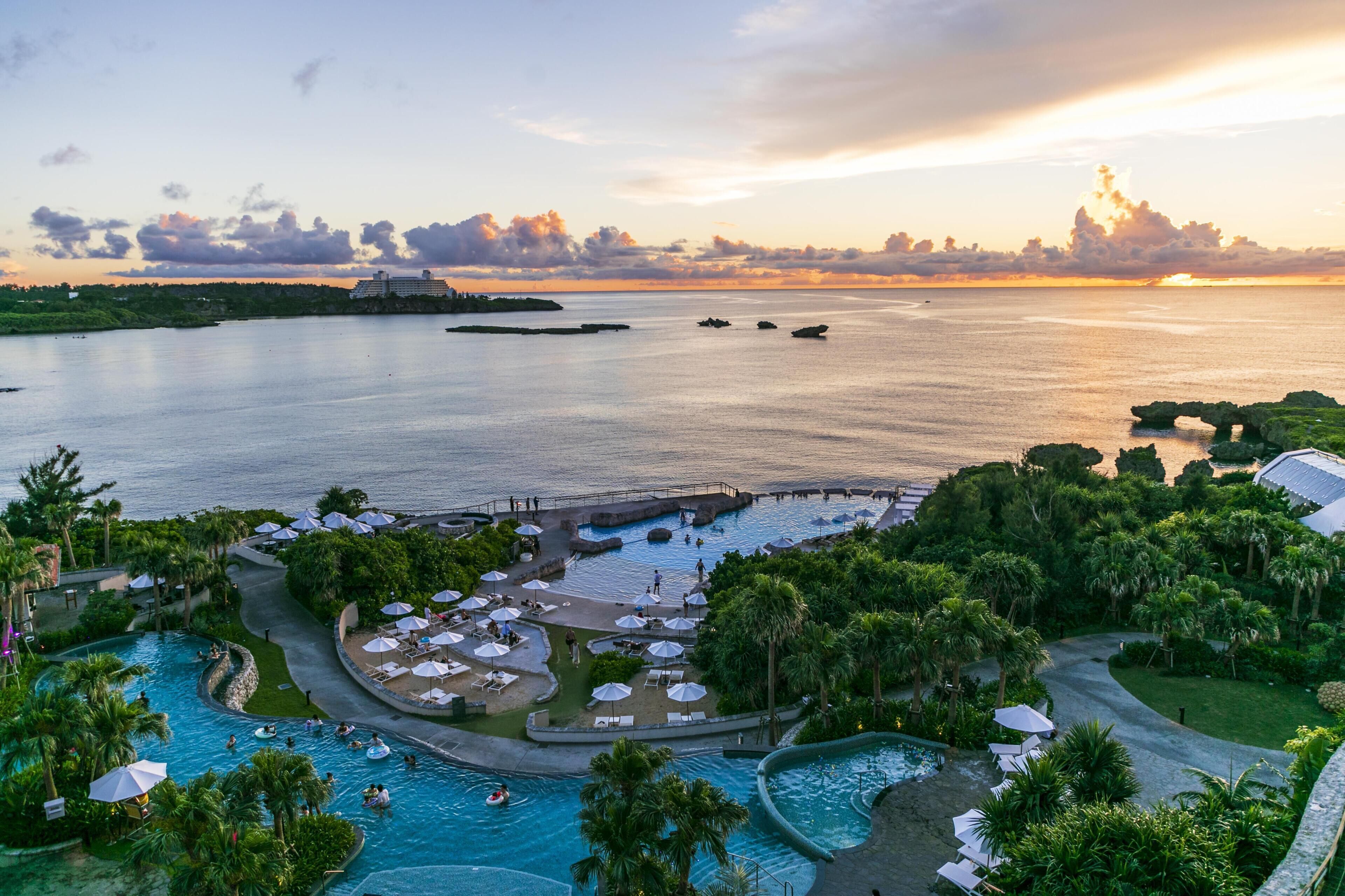 Un complexe hôtelier doté d’une grande piscine, de palmiers et offrant une vue sur la mer au coucher du soleil.