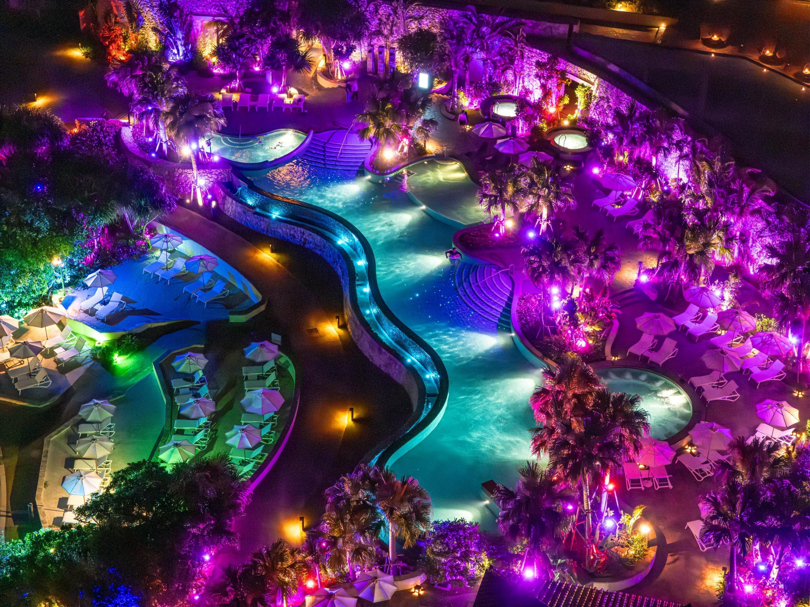 An aerial view of a resort with multiple swimming pools, lounge areas with umbrellas, and a well-lit landscape.