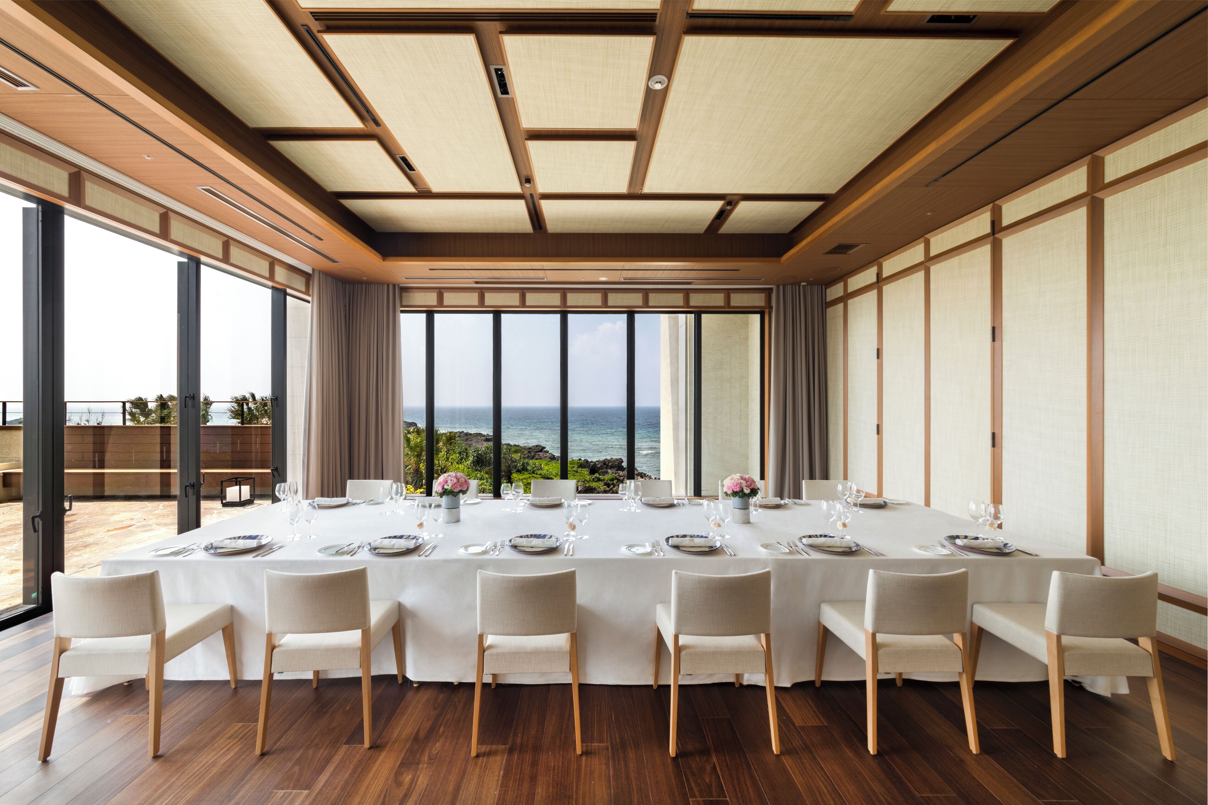 A dining room with a long table set for a meal, large windows with a view of the ocean, and a traditional wooden ceiling.