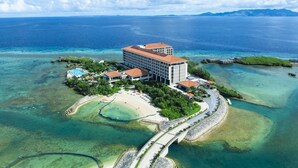 Vue aérienne d’un complexe hôtelier comprenant un grand bâtiment, plusieurs structures plus petites, une plage et des eaux d’un bleu limpide.