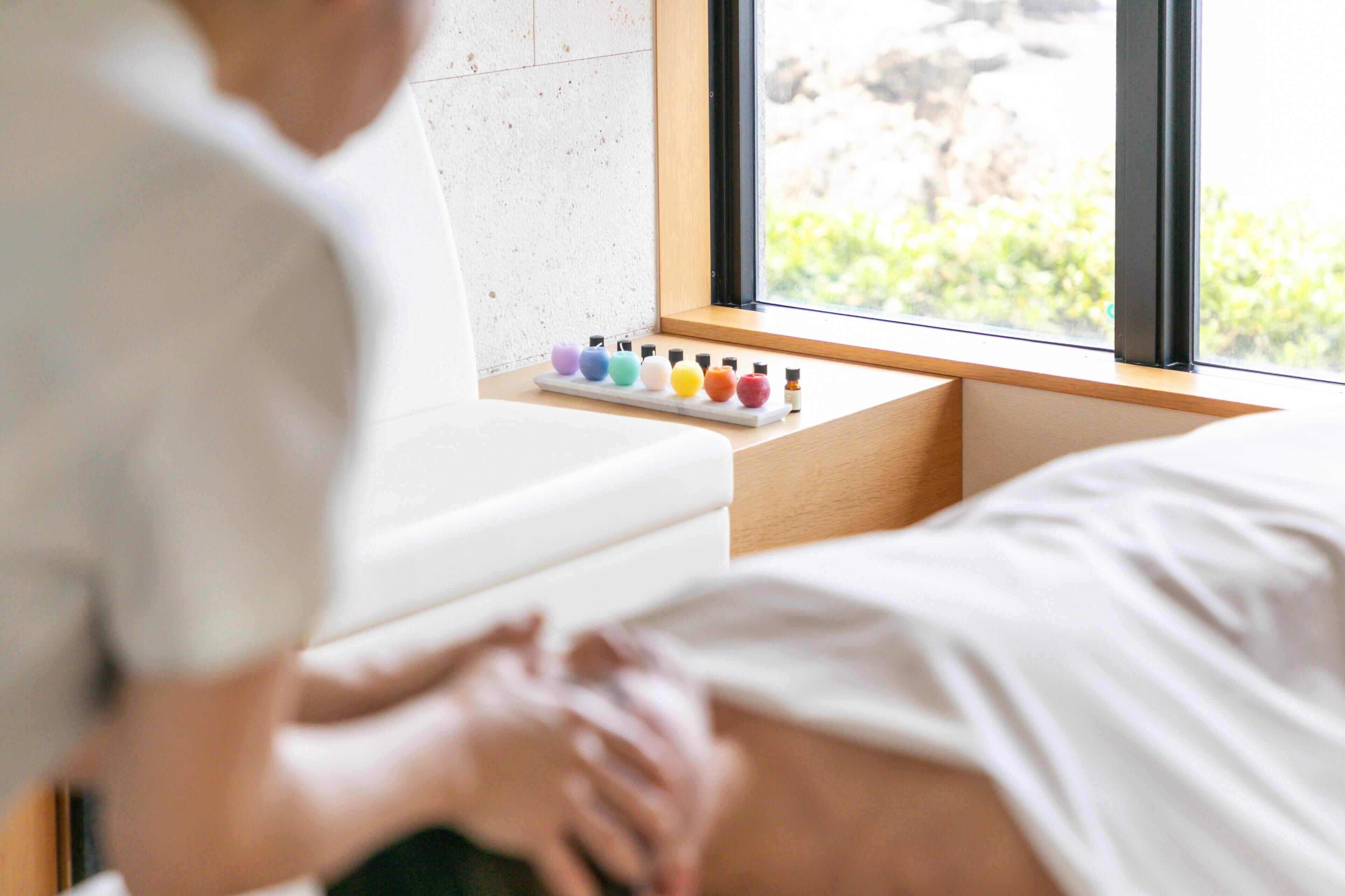 A person lying on a bed with a window view, a shelf with colorful bottles, and a white wall.