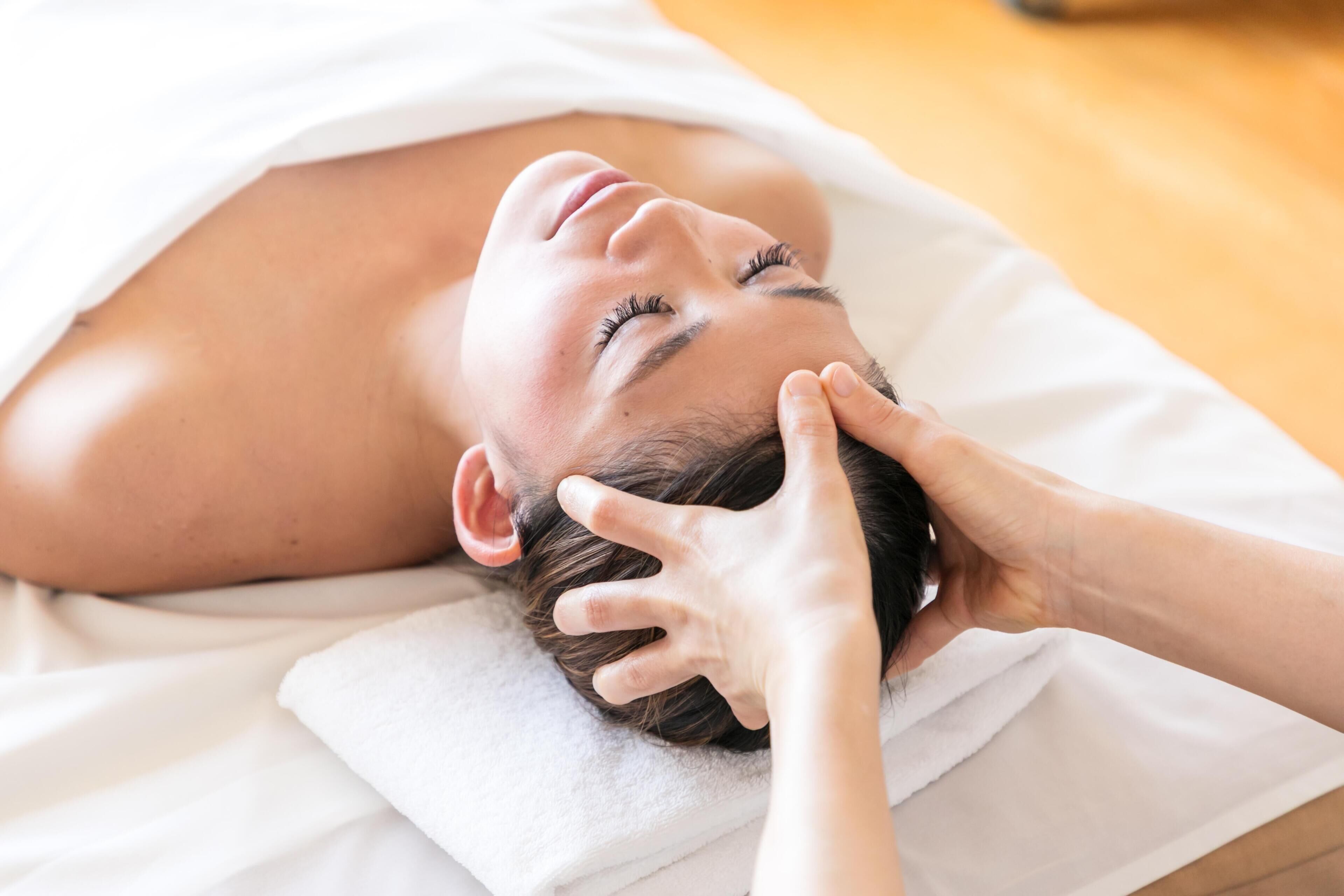 A person lying on a bed with a white towel, receiving a head massage.