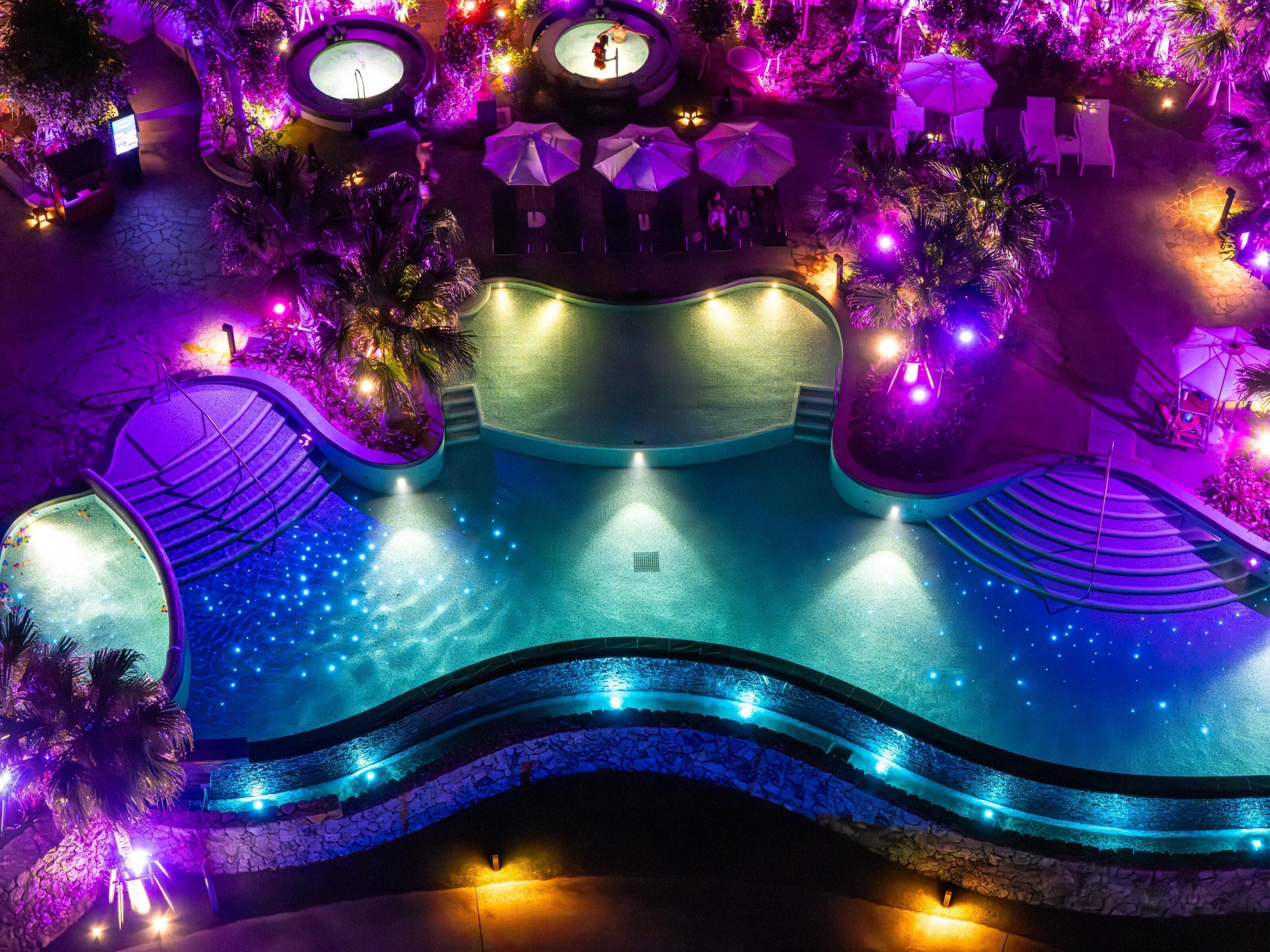 A pool area with a curved design, surrounded by palm trees and umbrellas, and illuminated by ambient lighting.