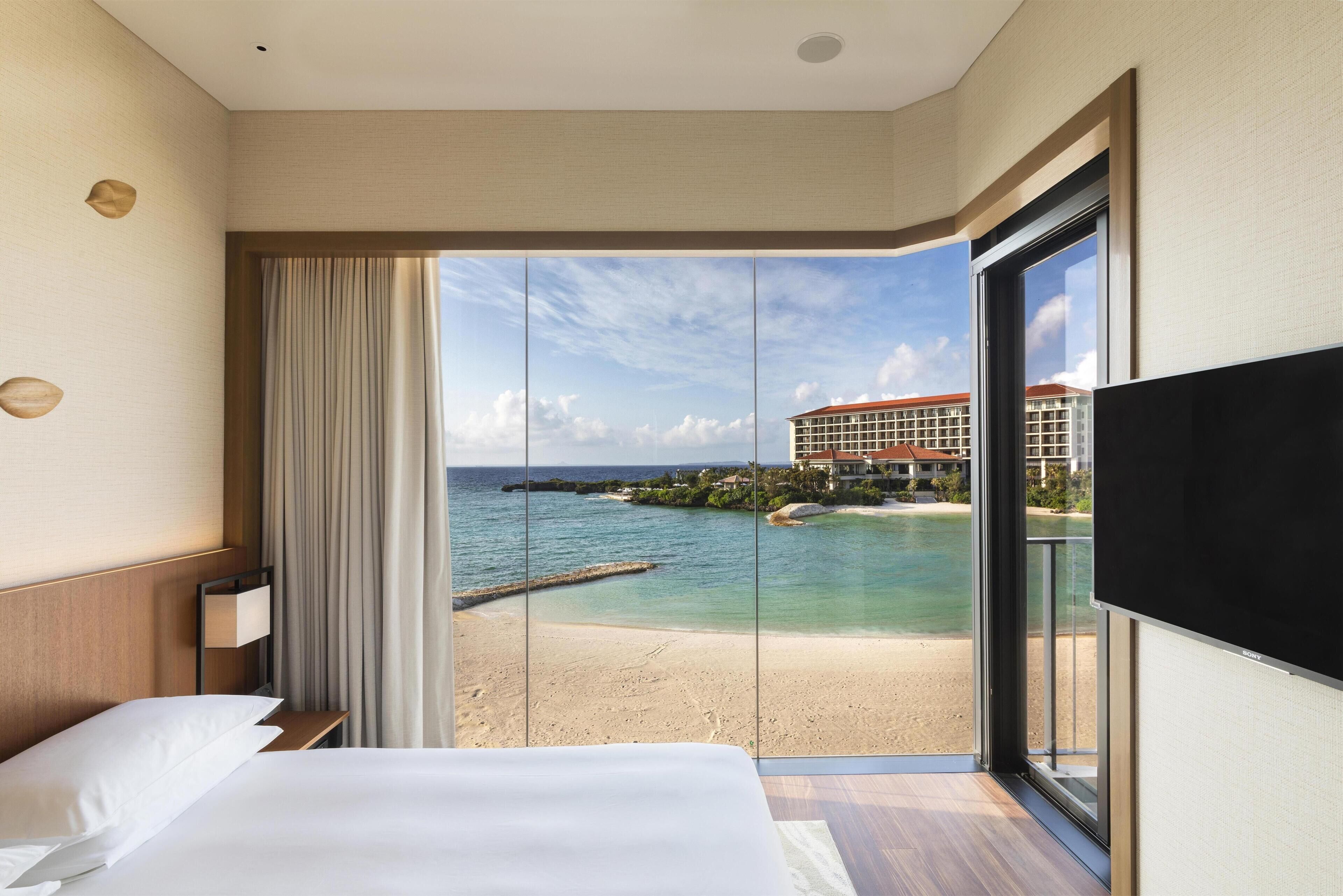 A hotel room with a bed, a TV, a window with a view of a beach and buildings.