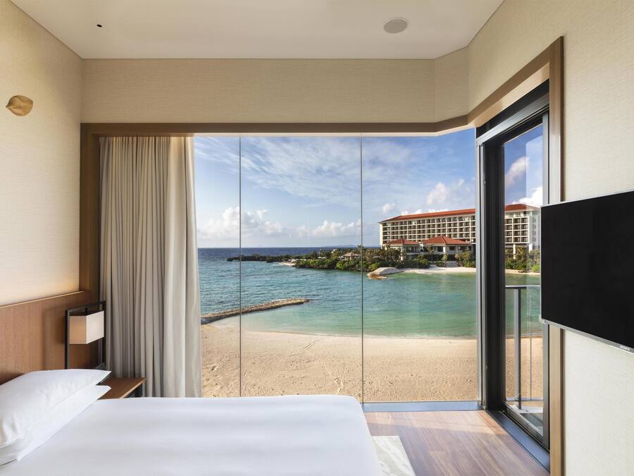 A hotel room with a bed, a TV, a window with a view of a beach and buildings.