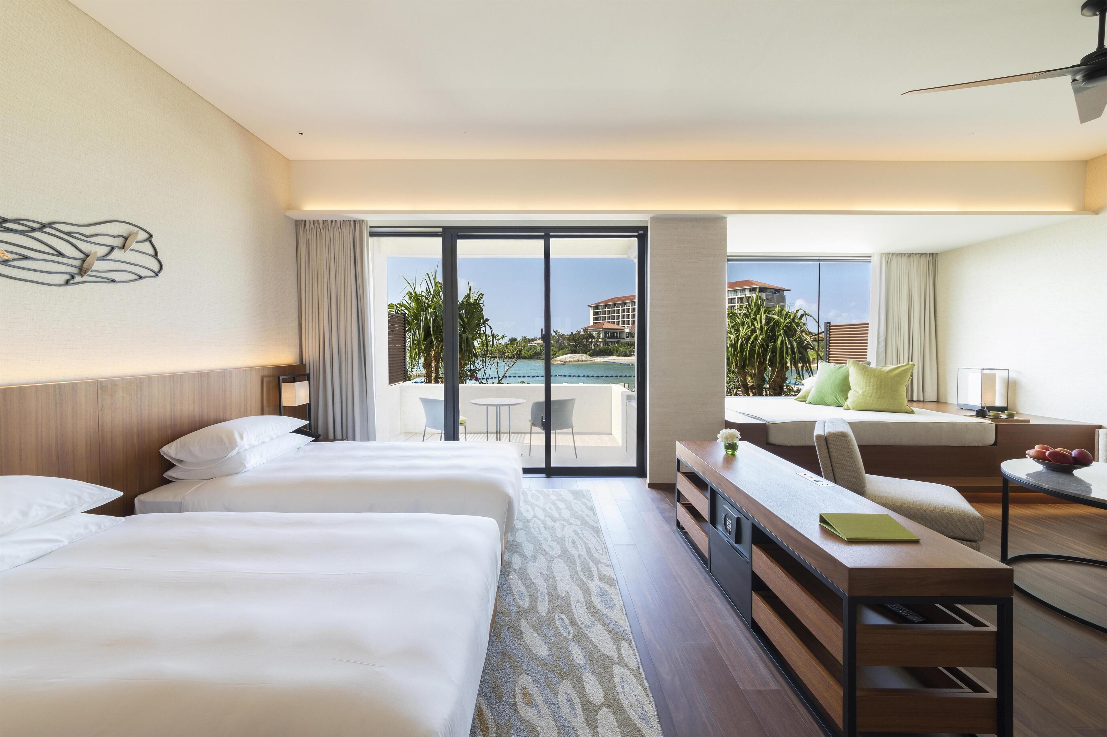 A hotel room with two beds, a desk, a chair, a small table, and a view of the pool and buildings.