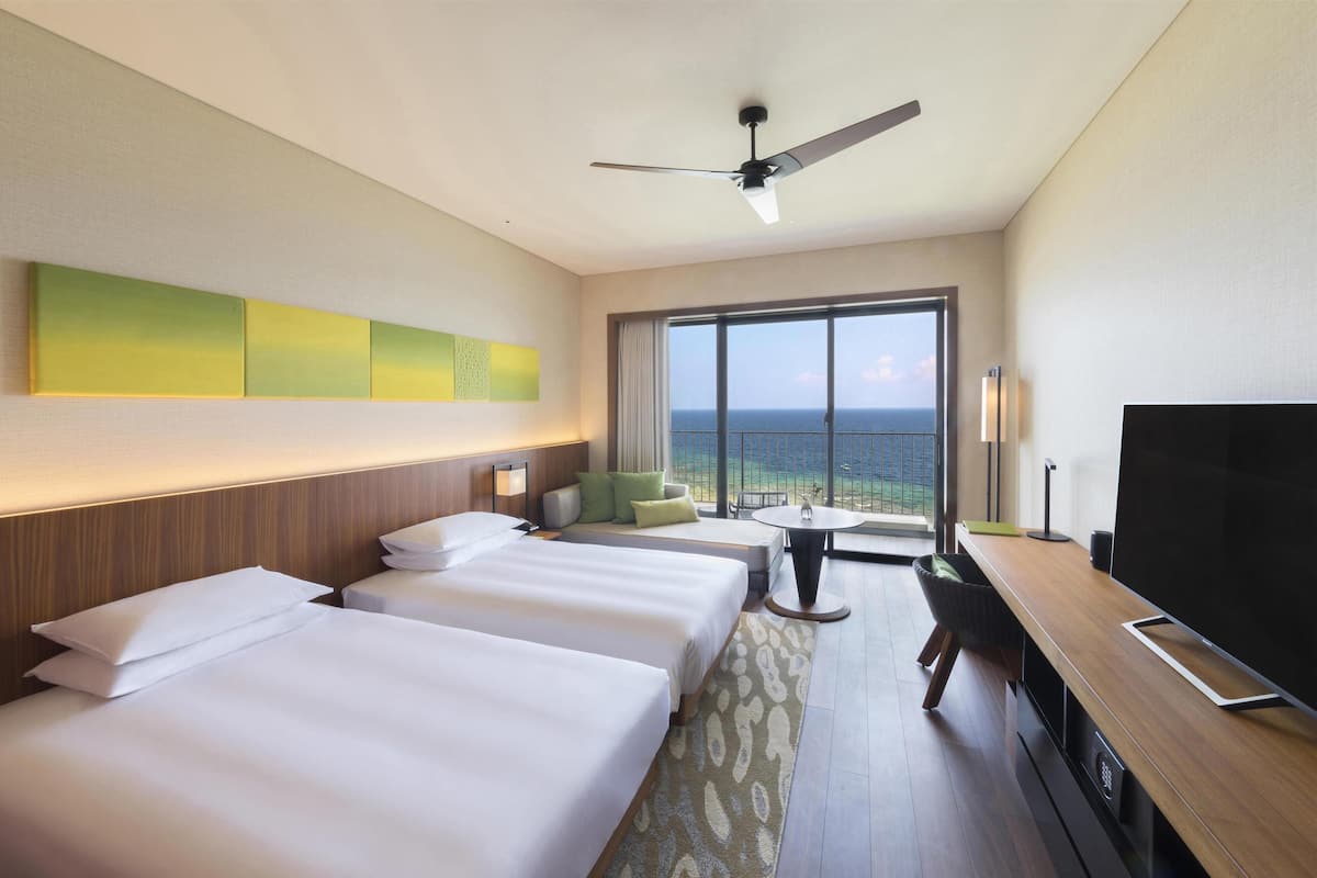 A hotel room with two beds, a TV, a ceiling fan, a window with a view of the sea, and a small table.