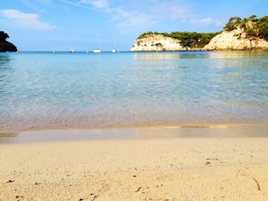 A beach with clear water, boats, and rocky cliffs.