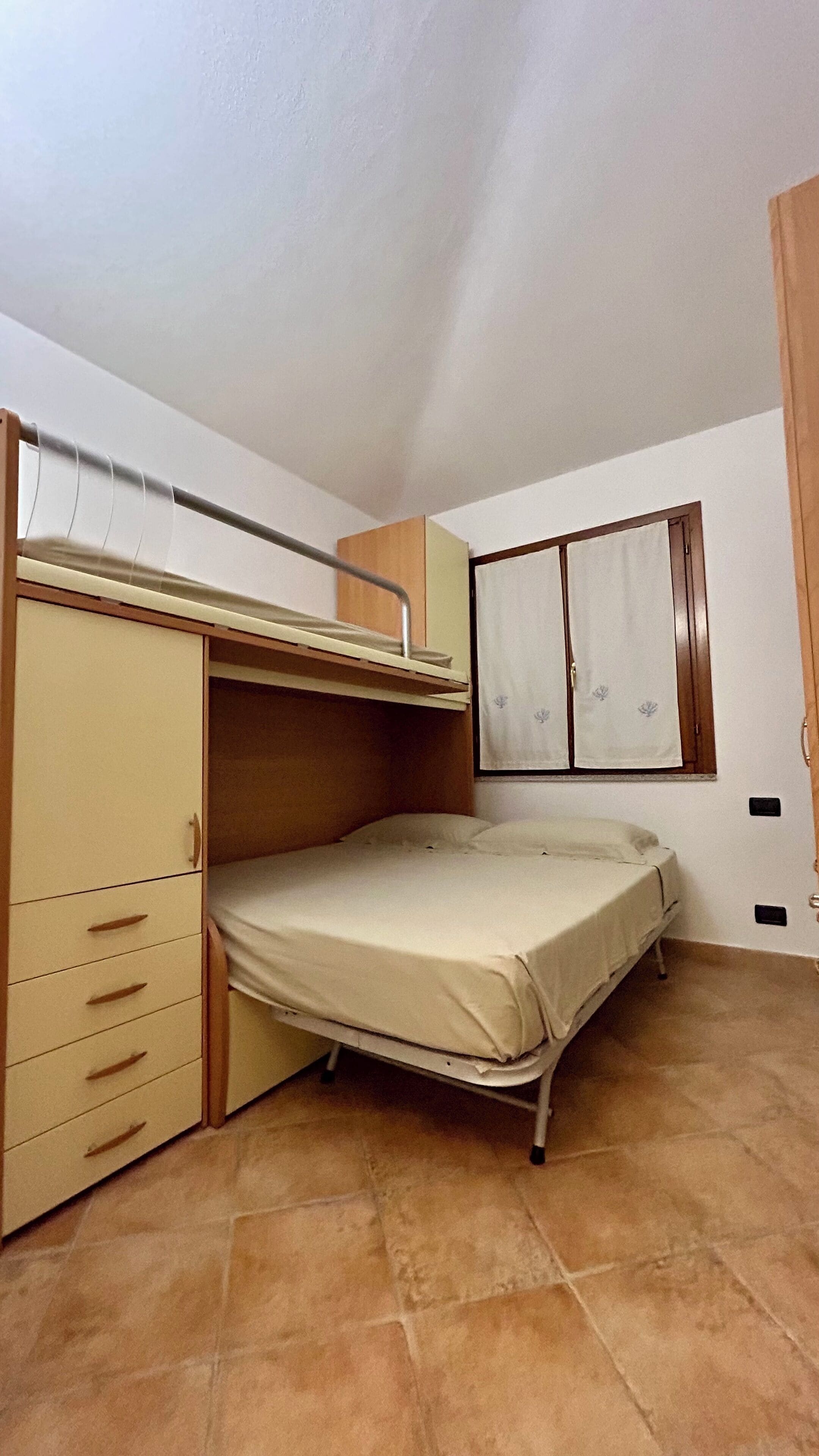 A compact room with a bunk bed, a wardrobe with multiple drawers, and a small window with curtains.