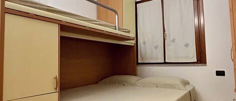 A compact room with a bunk bed, a wardrobe with multiple drawers, and a small window with curtains.